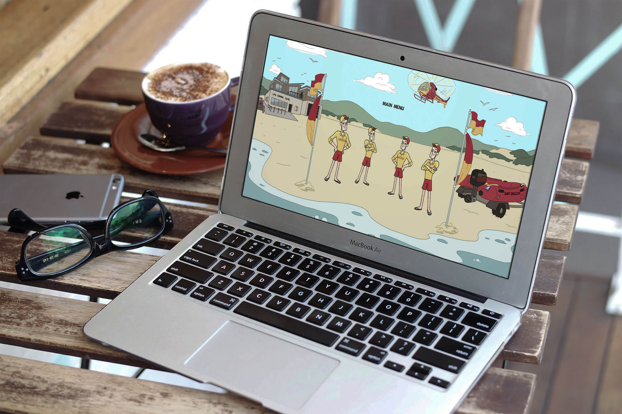 A laptop on a wooden table displays a cartoon beach scene with lifeguards, perfect for showcasing a creative portfolio. Nearby are a pair of glasses, an iPhone, and a cup of cappuccino on a saucer with a spoon.
