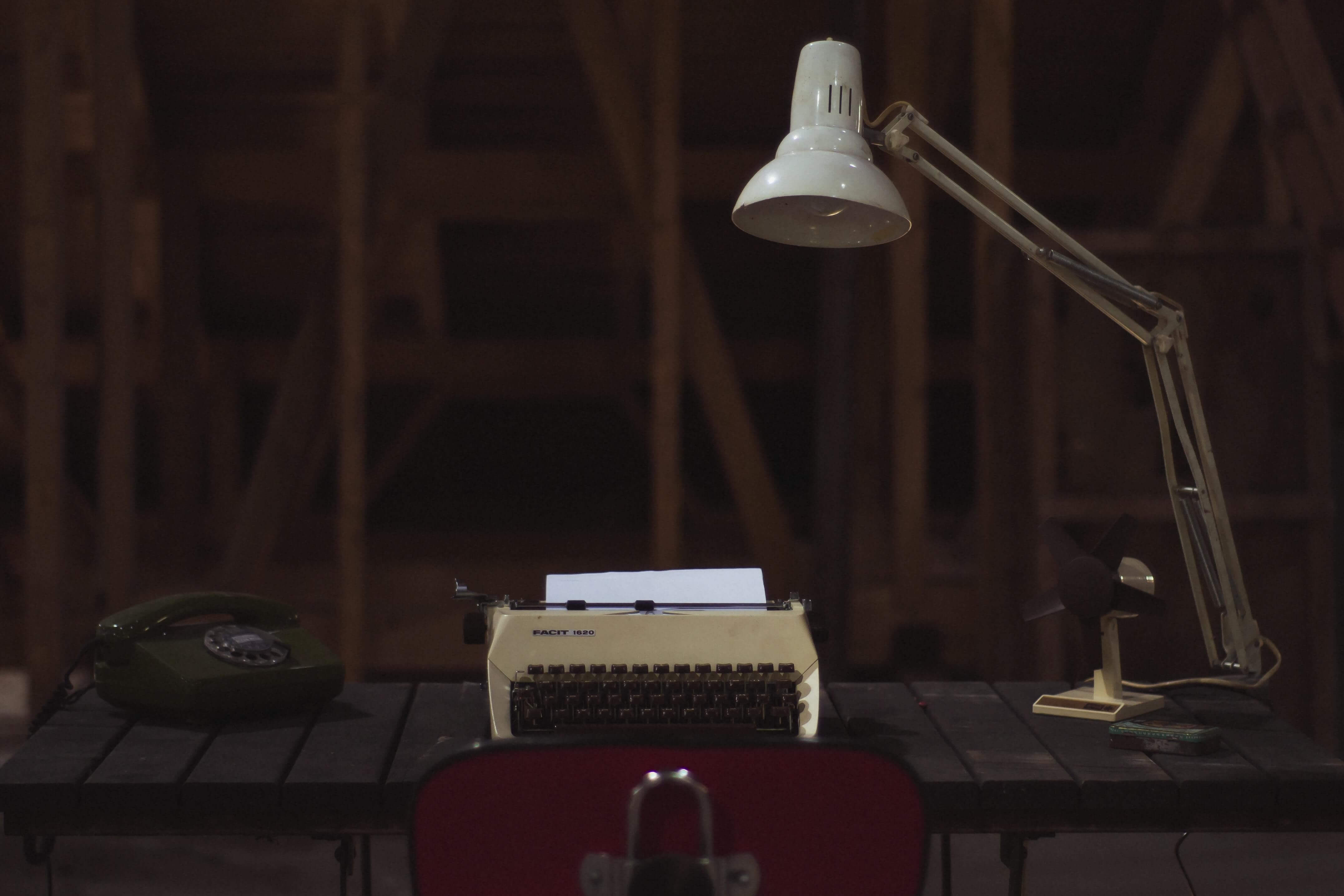 A vintage desk with a rotary telephone, typewriter with paper, desk lamp, and small fan, set against a dimly lit, wooden background.