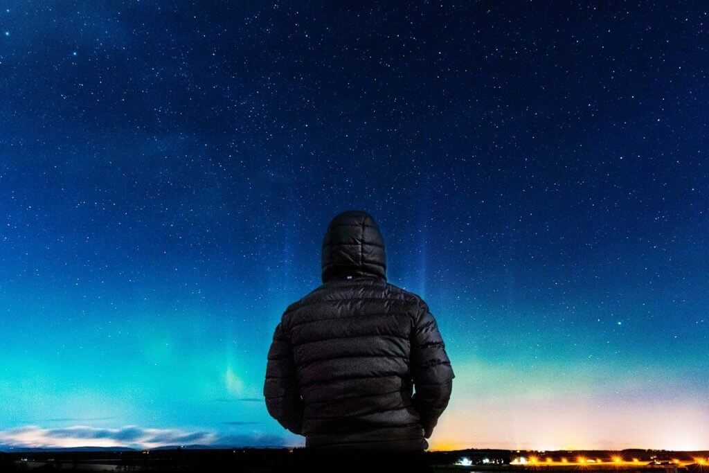 A person wearing a hooded jacket stands under a starry night sky, facing away, with faint northern lights and the glow of city lights on the horizon.