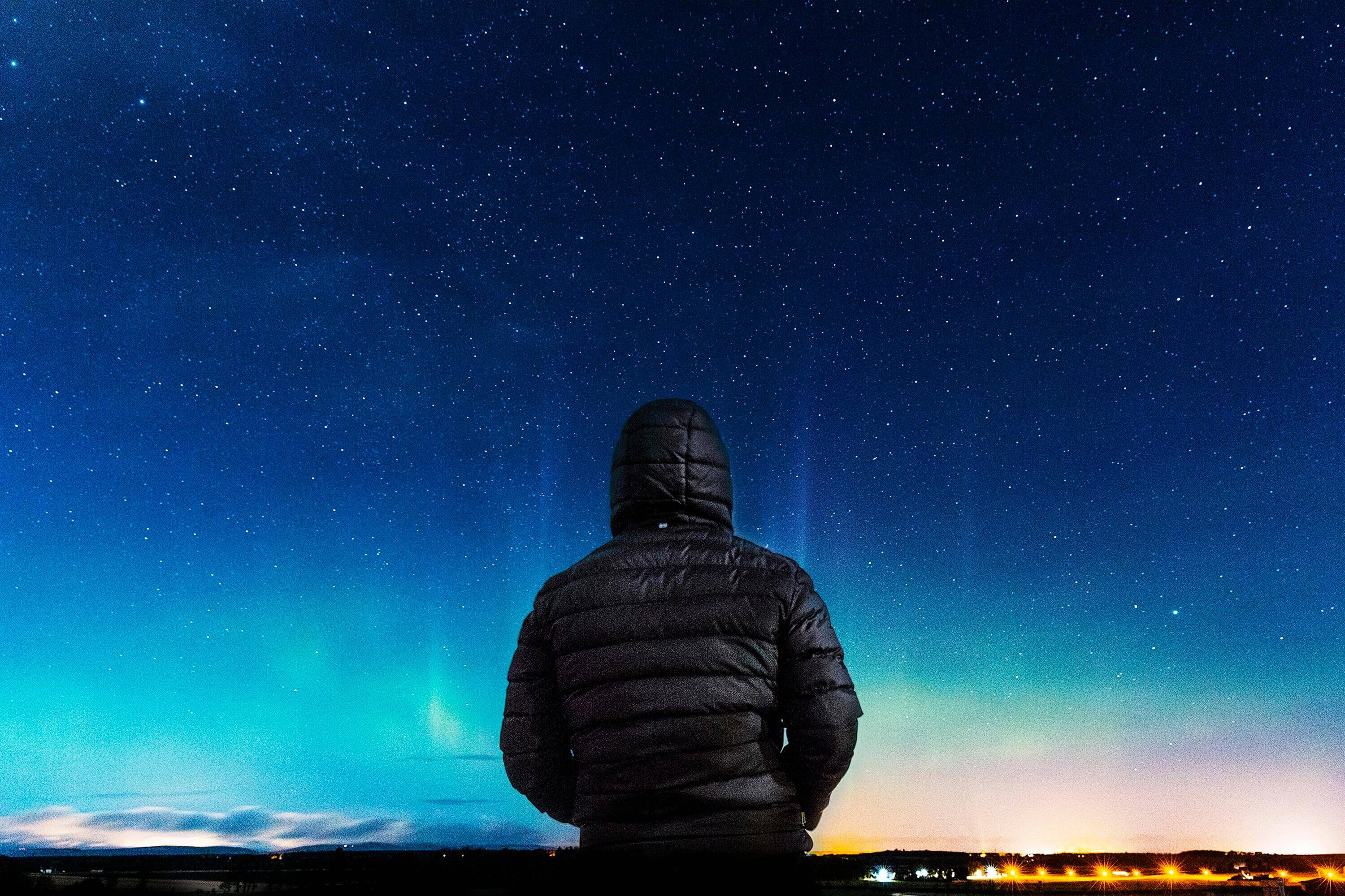 A person wearing a hooded jacket stands under a starry night sky, facing away, with faint northern lights and the glow of city lights on the horizon.