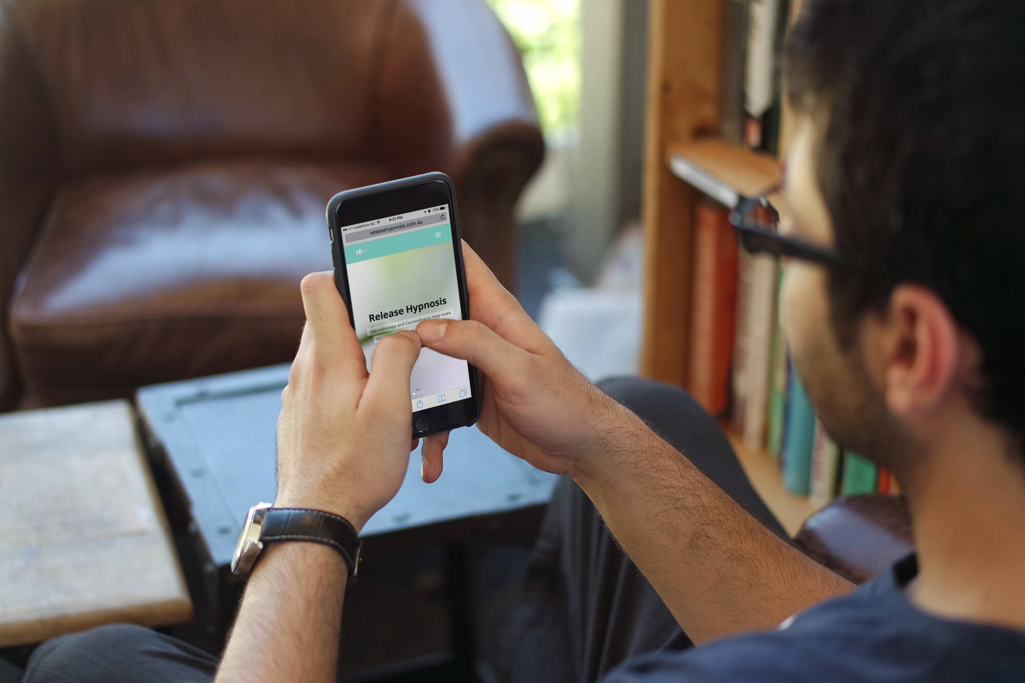 A person wearing glasses sits in a cosy room and uses a mobile phone. The phone screen displays a webpage with the heading “Release Hypnosis.” Books and a leather chair are visible in the background.