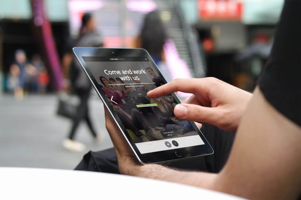A person adapts to urban life, holding and using a tablet outdoors. On the screen, a website displays "Come and work with us," with blurred city surroundings in the background.