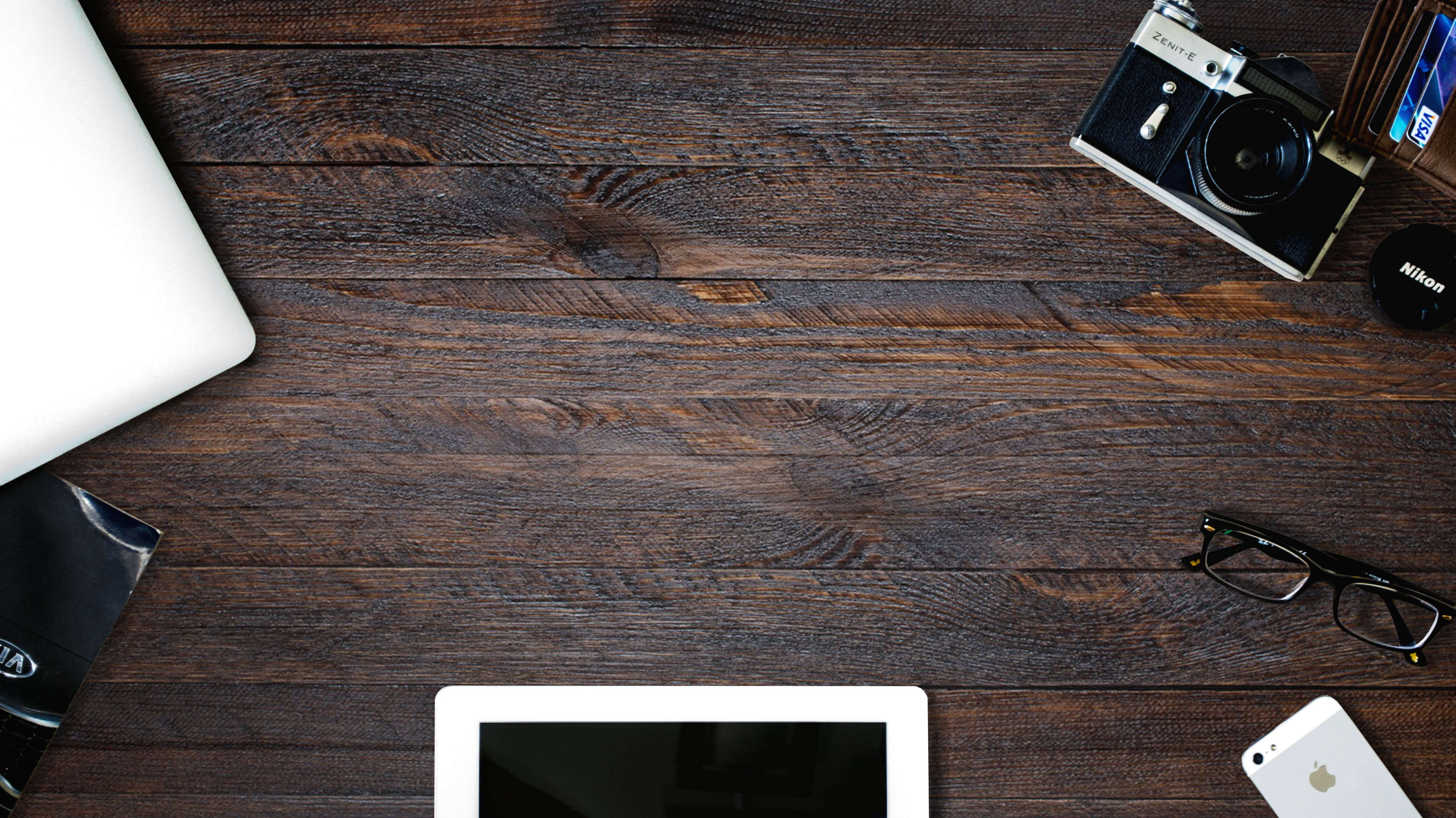 A dark wooden desk, adapted for creativity, with a camera, lens cap, glasses, mobile phone, part of a laptop, a magazine, and the edge of a tablet with a black screen arranged around the corners.