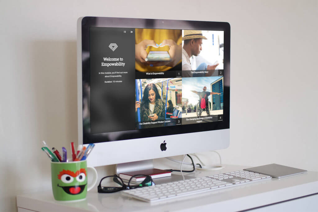 An iMac on a white desk displays a website about Empowerability and disability support training, featuring welcome text and images of diverse people. The desk holds office supplies, a keyboard, mouse, mobile phone, and a mug with a face.