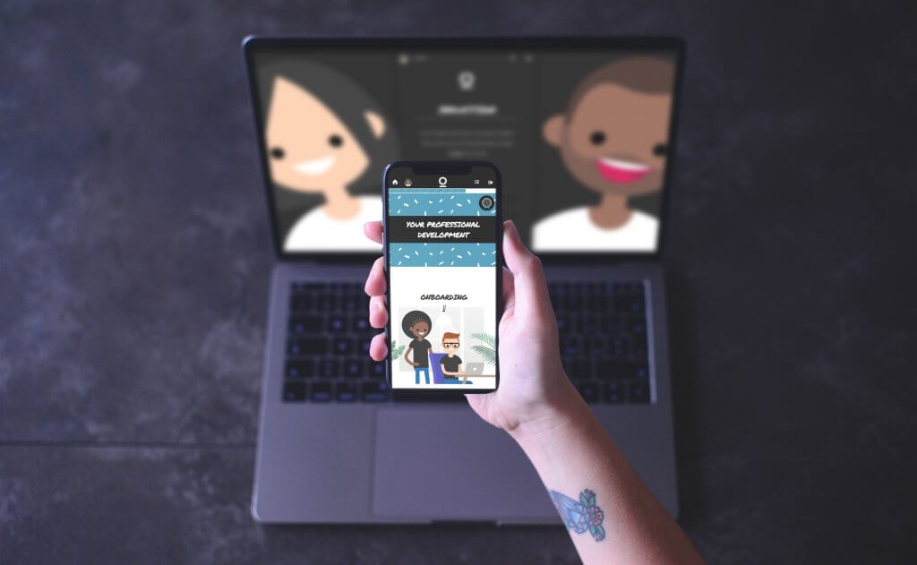 A person holds a mobile phone displaying a cartoon image, with a laptop in the background showing two cartoon faces on its screen—perhaps winners of cartoon awards—all set on a dark surface.