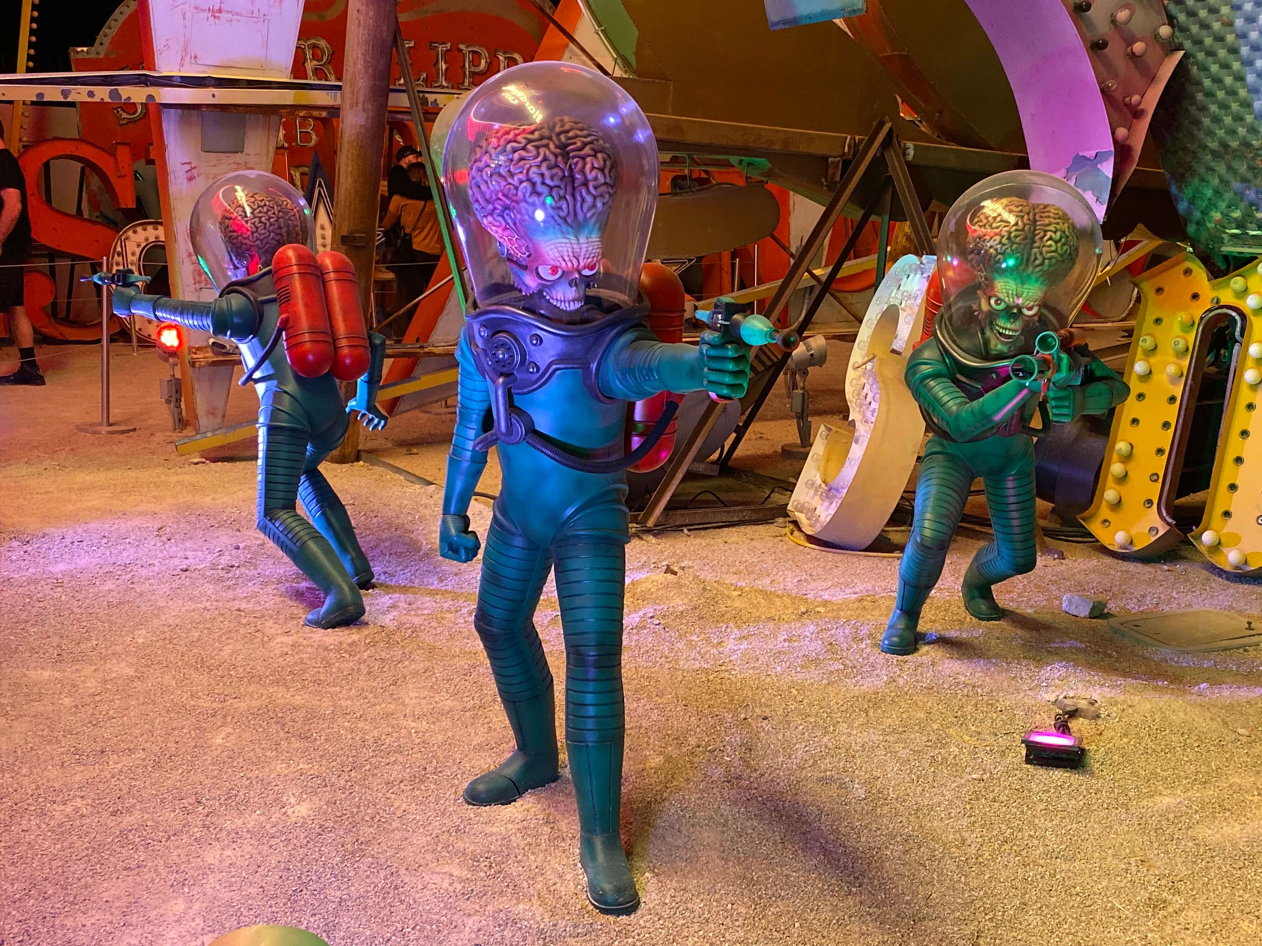 Three figures in green spacesuits with oversized brains stand on a colourful, sci-fi DevLearn set, aiming ray guns in different directions as bright lights and neon signs glow in the background.