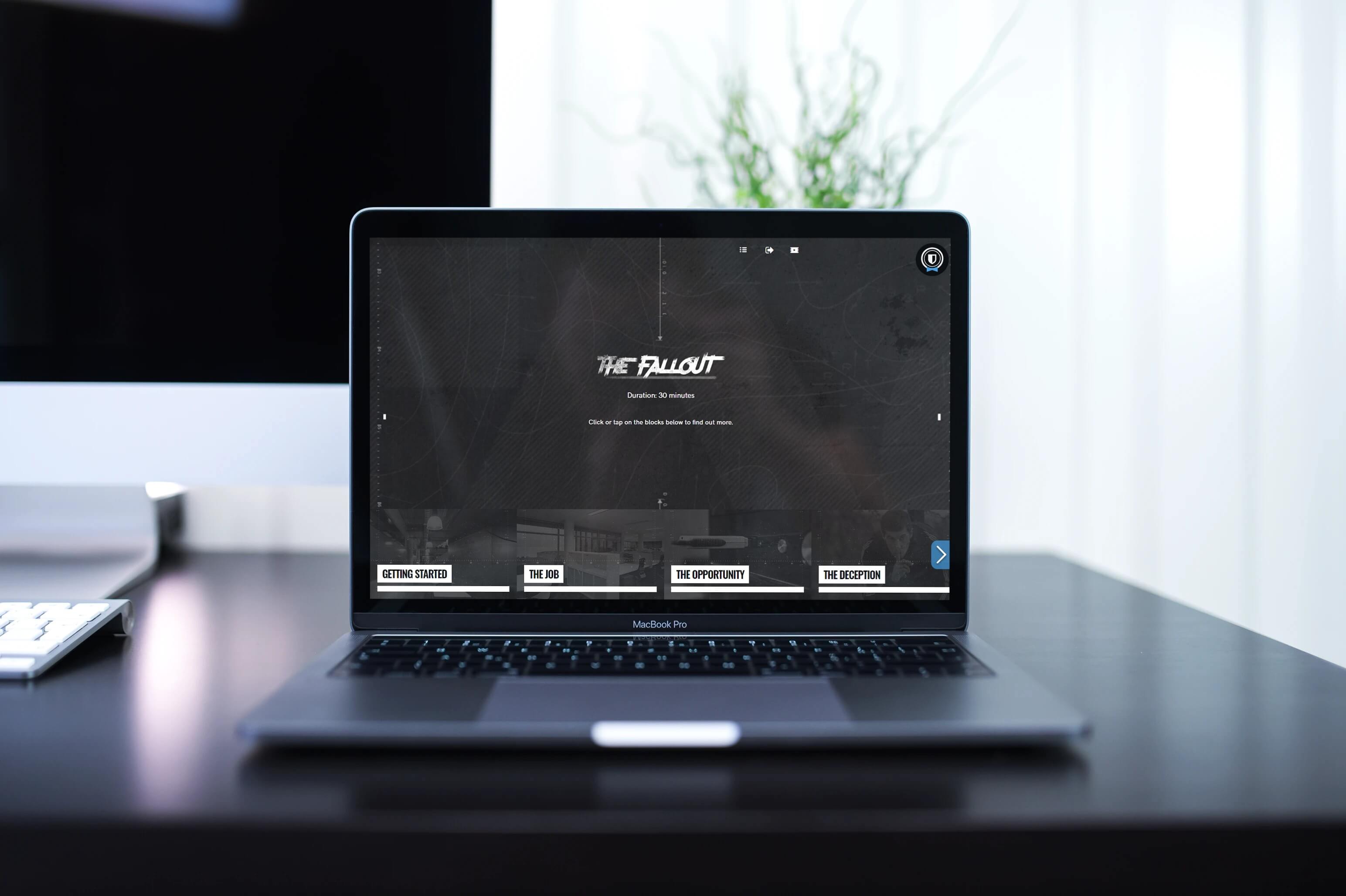 A MacBook Pro on a dark desk displays a website titled "The Fallout," highlighting its recent awards. The background is blurred with a keyboard, monitor, and a green plant visible. The scene is bright and modern.