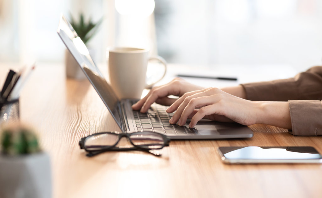 Close-up of a person typing on a laptop at a wooden desk, surrounded by items that highlight the value of design—a mug, mobile phone, spectacles, and small plant—all set in a bright, softly lit environment.